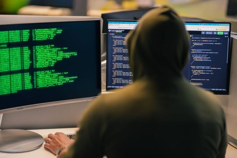 A person in a hoodie coding on dual monitors, depicting cybersecurity and hacking themes.
