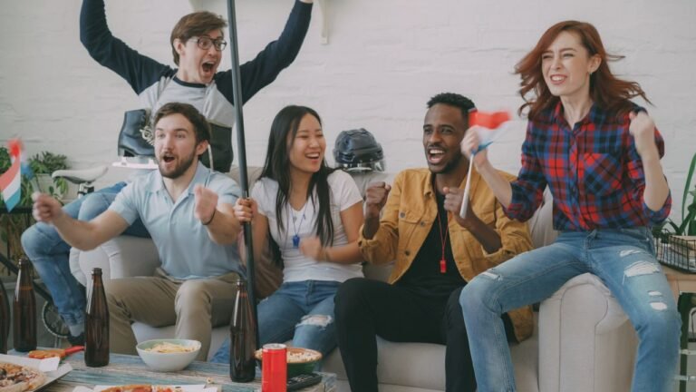 Group of diverse friends cheering indoors, holding flags, enjoying a sports victory celebration.