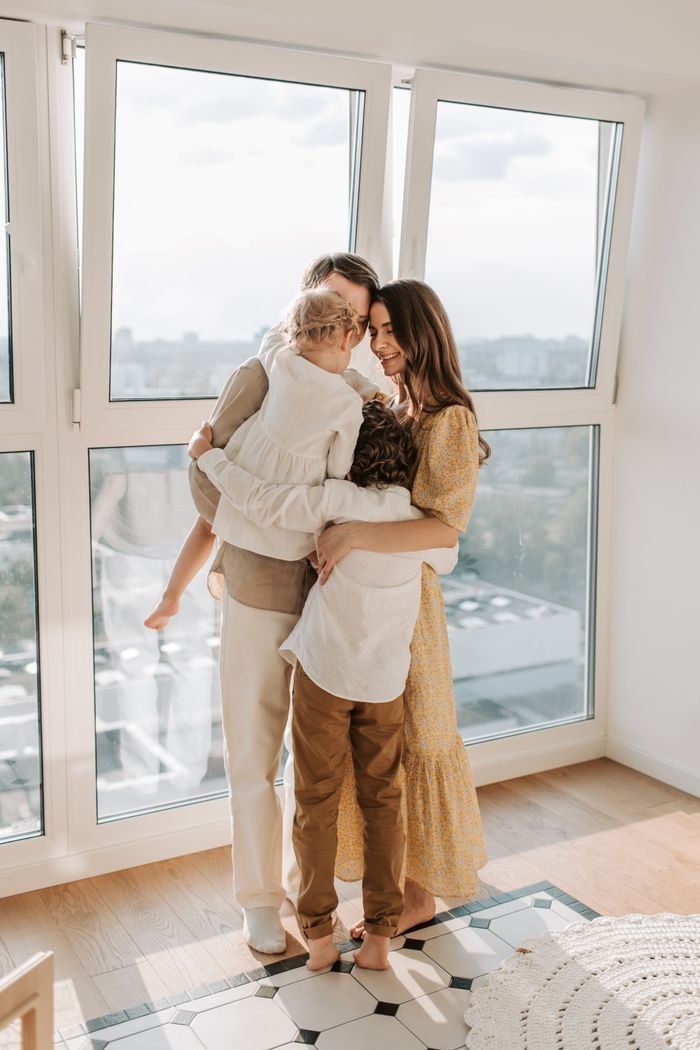 Heartwarming family group hug indoors, sunlight filtering through large windows with a city backdrop, illustrating the family support that matters in a family emergency