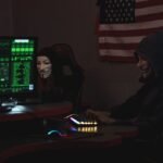 Two anonymous individuals wearing hacker masks use computers in a dark room with an American flag backdrop, highlighting cyber security themes.