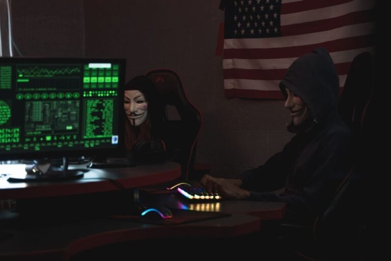 Two anonymous individuals wearing hacker masks use computers in a dark room with an American flag backdrop, highlighting cyber security themes.