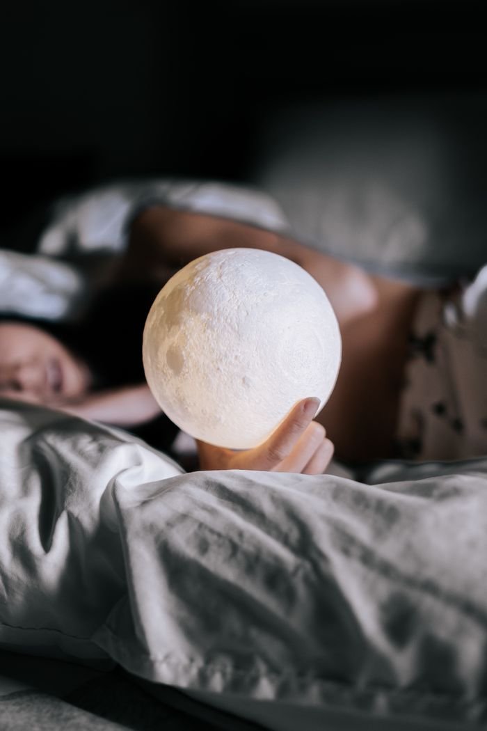 A woman lying in bed holding a glowing moon lamp in a cozy, dimly-lit setting.