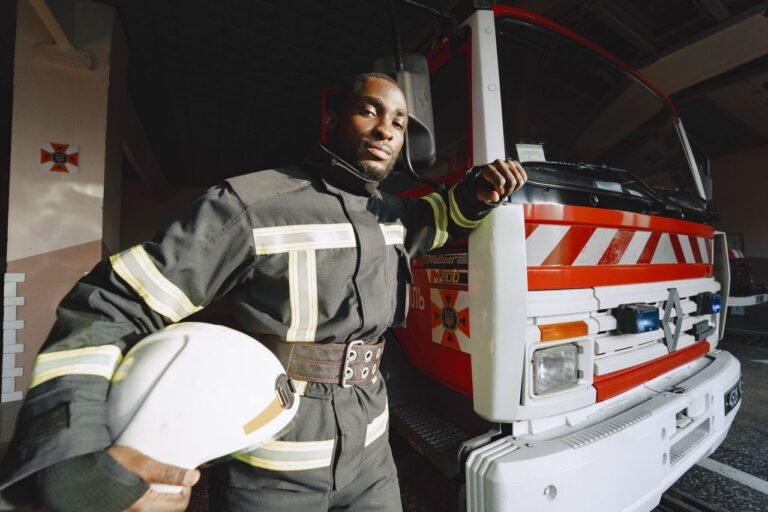 Firefighter in uniform poses confidently beside a fire truck indoors.